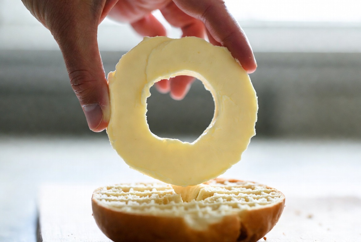 Hand placing full bagel-sized Butter Ring or Cream Cheese Ring onto a scored bagel showing perfect coverage